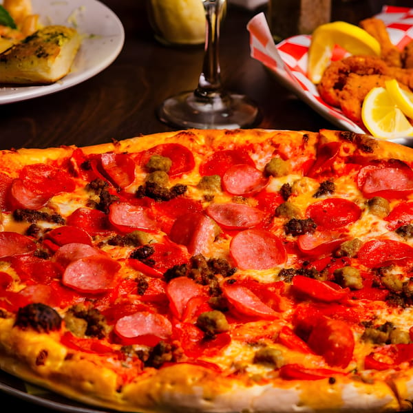 A close-up of a pepperoni and sausage pizza with other dishes blurred in the background.