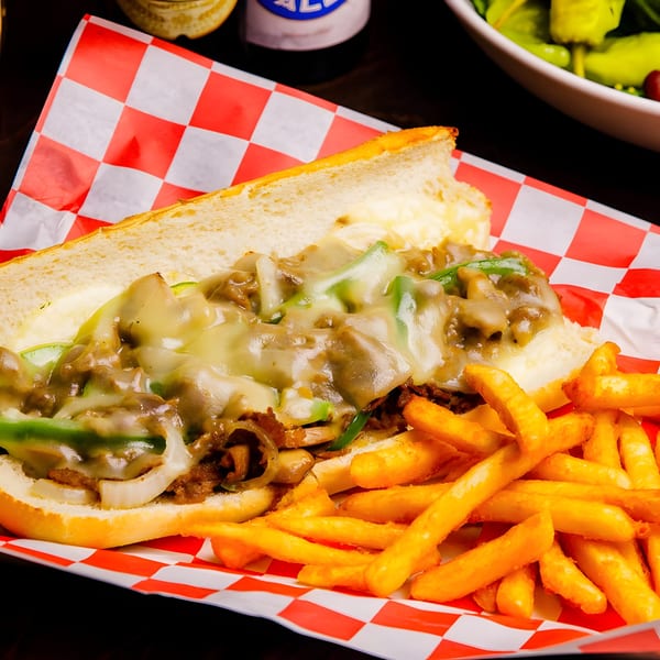 Cheesesteak sandwich with melted cheese, peppers, and onions, served with golden fries on checkered paper.