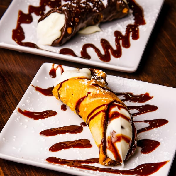 Two cannolis with white cream filling, drizzled with chocolate sauce and powdered sugar on white plates.