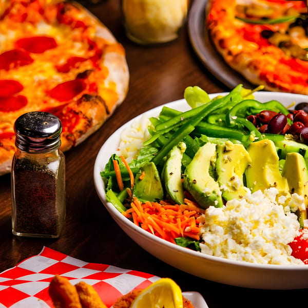 A vibrant salad with avocado, feta, and bell peppers, alongside pizzas and a pepper shaker.