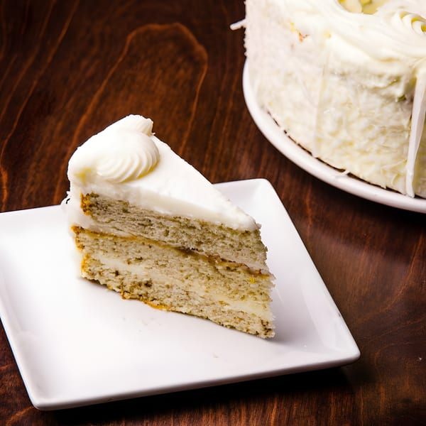 A slice of white cake with white frosting on a white plate, with a whole cake in the background.