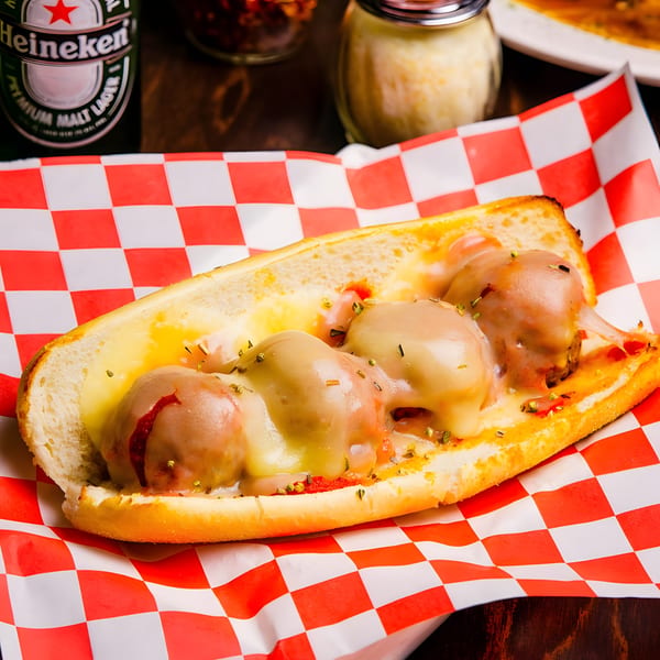 Meatball sub with melted cheese and herbs on red and white checkered paper, with a beer bottle in the background.